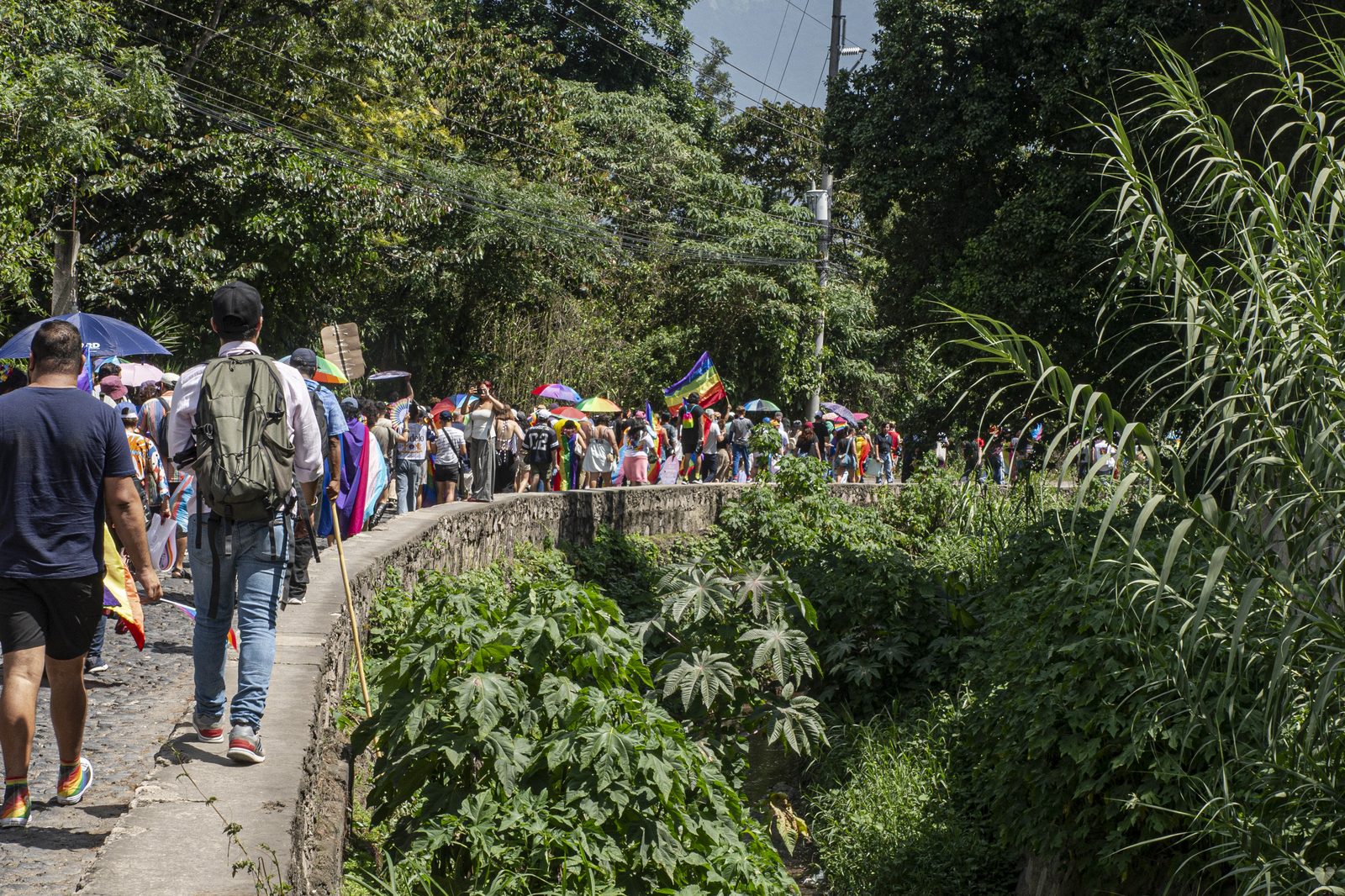 Personas caminando junto a un canal con banderas arcoíris y el volcán al fondo